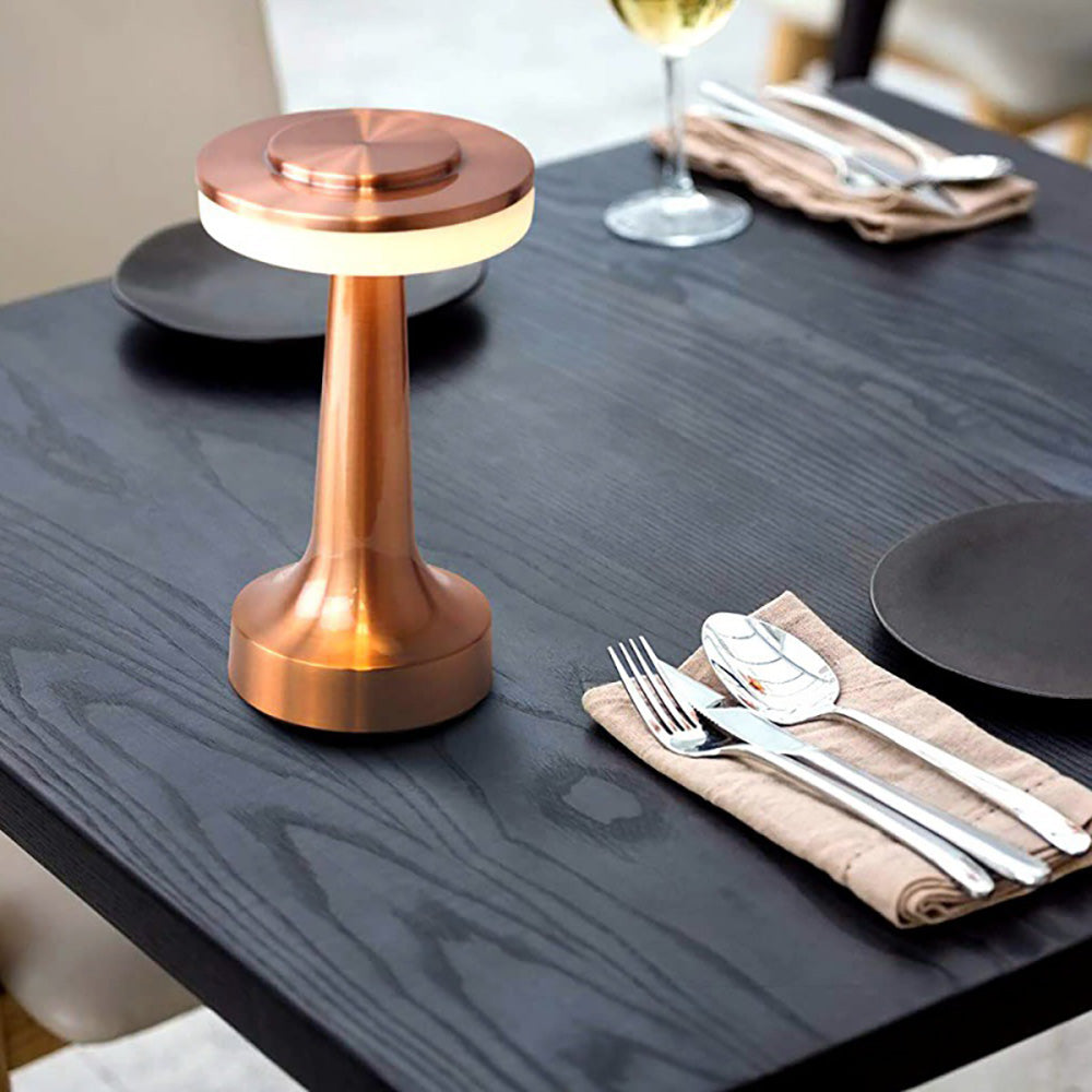 Lampe de table en cuivre moderne sur une table en bois sombre, entourée de couverts et assiettes, créant une ambiance élégante et sophistiquée.