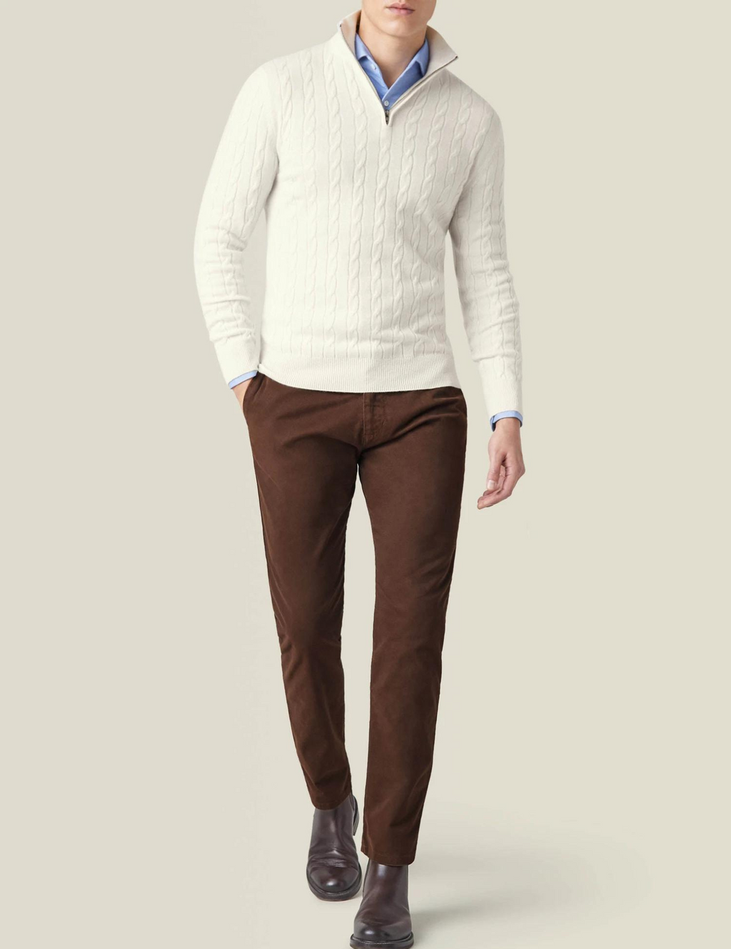 Homme portant un pull blanc en maille torsadée, chemise bleue, pantalon marron et bottines en cuir. Mode masculine élégante et décontractée.