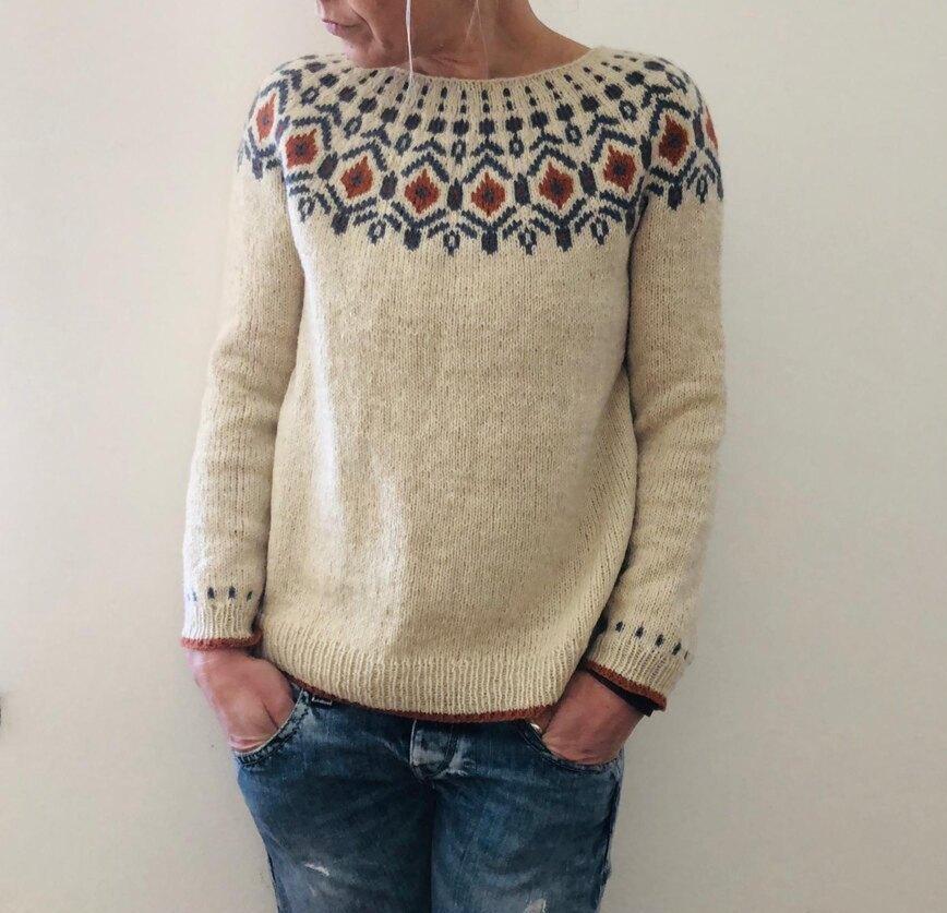 Pull en laine beige avec motif nordique coloré, manches longues, porté par une personne en jean. Mode hiver, tricot tendance, style décontracté.
