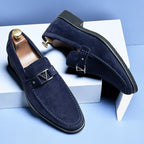 Chaussures mocassins en daim bleu marine pour homme avec boucle métallique, posées sur fond bleu et blanc. Élégance, mode masculine, chaussures de luxe.
