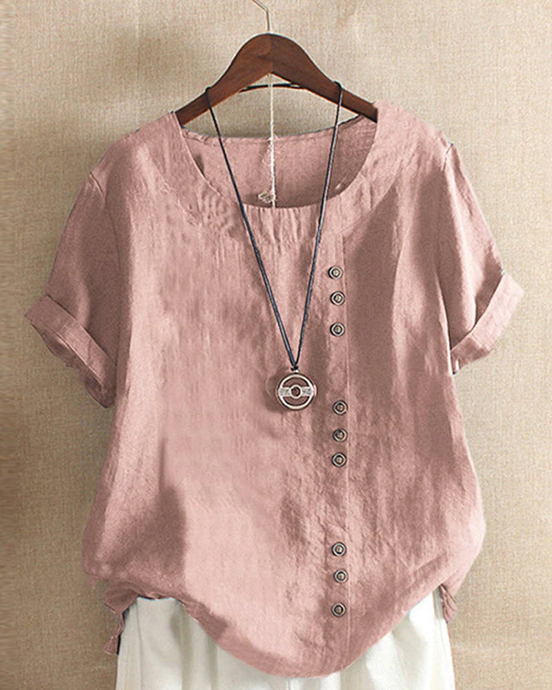 Blouse en lin rose avec boutons asymétriques, manches courtes retroussées, col rond, suspendue sur cintre, collier pendentif en bois.