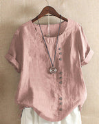 Blouse en lin rose avec boutons asymétriques, manches courtes retroussées, col rond, suspendue sur cintre, collier pendentif en bois.