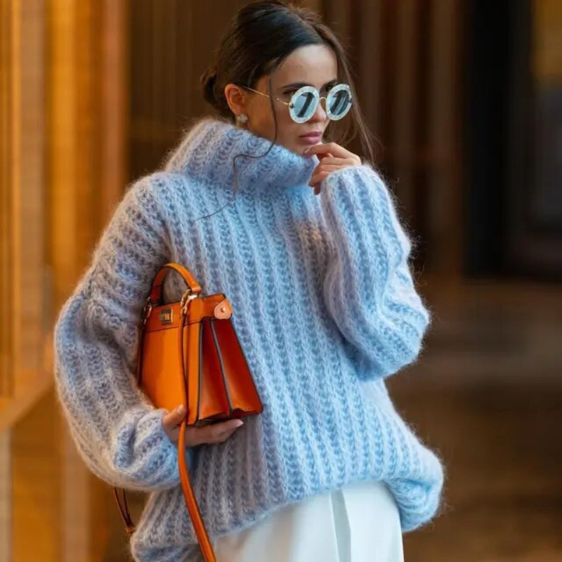 Femme élégante portant un pull en laine bleu, lunettes de soleil rondes et sac à main orange vif, mode automne-hiver chic et tendance.