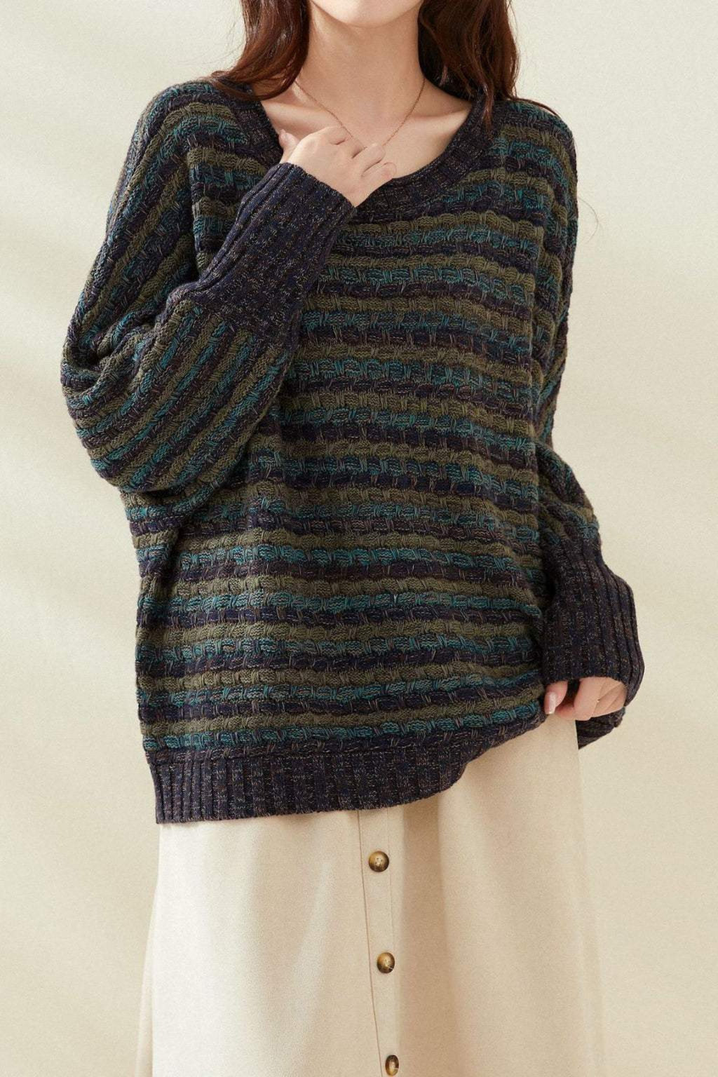 Femme portant un pull rayé bleu et vert, manches longues, col en V, avec une jupe beige à boutons. Mode automne-hiver, style décontracté.