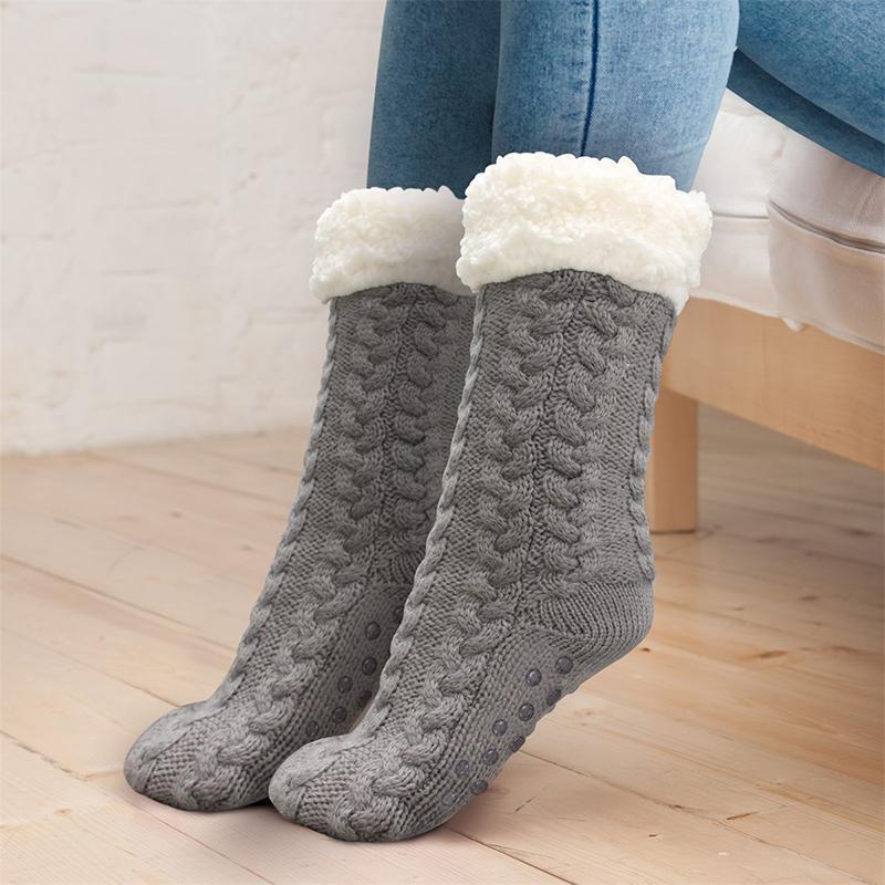 Chaussettes en laine grise avec doublure en fausse fourrure blanche, motif torsadé, portées par une personne assise sur un sol en bois clair.