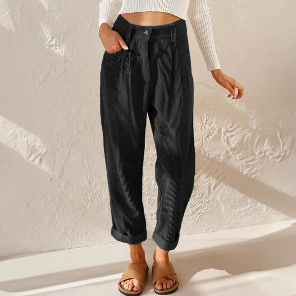 Femme portant un pantalon noir ample en velours côtelé, associé à un pull blanc à manches longues et des sandales marron, sur fond de mur texturé.