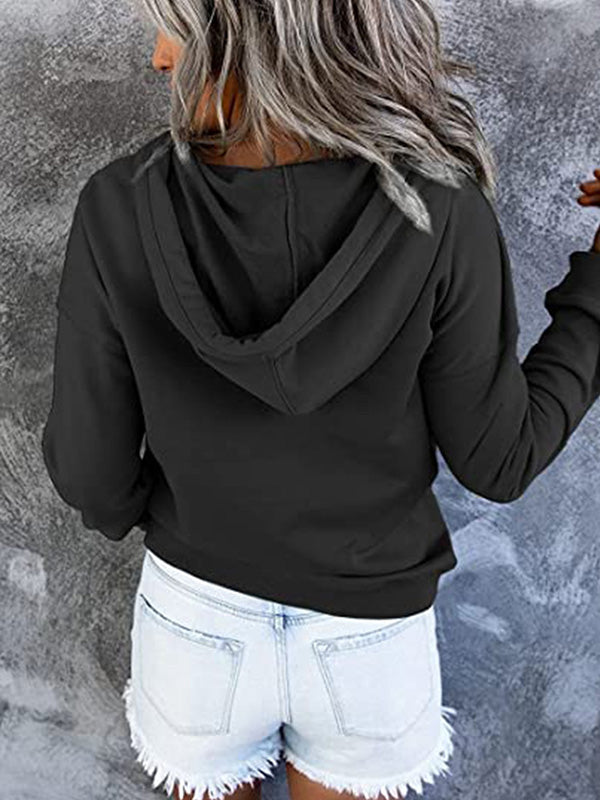 Femme portant un sweat à capuche noir et un short en jean délavé, posant de dos contre un mur gris texturé. Mode décontractée, style urbain.
