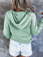 Femme de dos portant un sweat à capuche vert menthe et un short en jean déchiré, posant devant un mur gris texturé. Mode décontractée tendance.