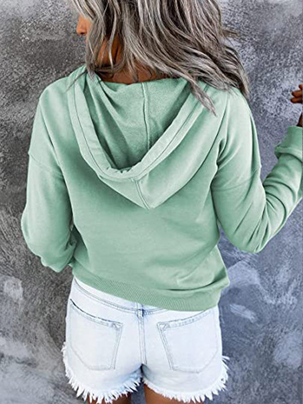 Femme de dos portant un sweat à capuche vert menthe et un short en jean déchiré, posant devant un mur gris texturé. Mode décontractée tendance.