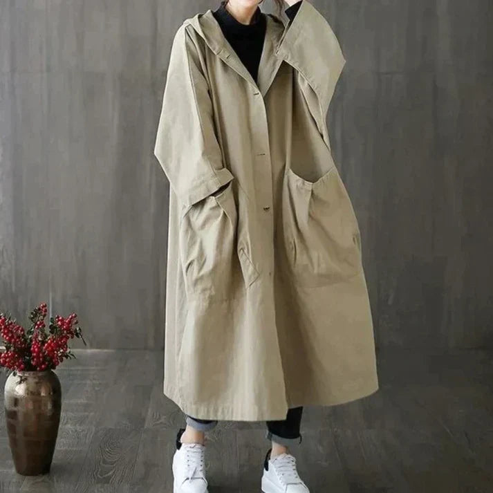 Manteau long beige à capuche pour femme, style ample et décontracté, poches avant, porté avec des baskets blanches, fond gris moderne.