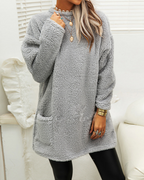 Femme portant un pull en peluche gris, manches longues, col rond, avec poches. Style décontracté, idéal pour l'hiver. Accessoires dorés visibles.