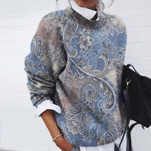 Femme portant un pull à motifs cachemire bleu et beige, manches longues, avec chemise blanche en dessous, tenant un sac noir. Mode automne chic.