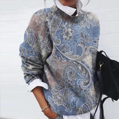 Femme portant un pull à motifs cachemire bleu et beige, manches longues, avec chemise blanche en dessous, tenant un sac noir. Mode automne chic.