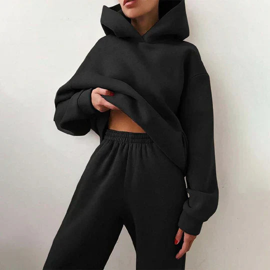Femme portant un ensemble de survêtement noir, composé d'un sweat à capuche ample et d'un pantalon de jogging, style décontracté et moderne.