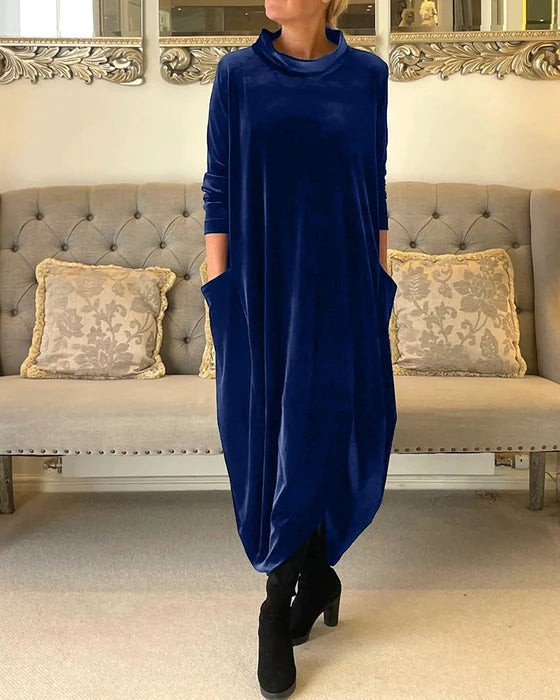 Robe longue en velours bleu avec poches, portée par une femme debout devant un canapé beige élégant, décoré de coussins à motifs.