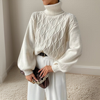 Femme portant un pull torsadé blanc à col roulé, tenant un sac à main marron avec chaîne dorée, sur fond de mur beige et tapis crème.