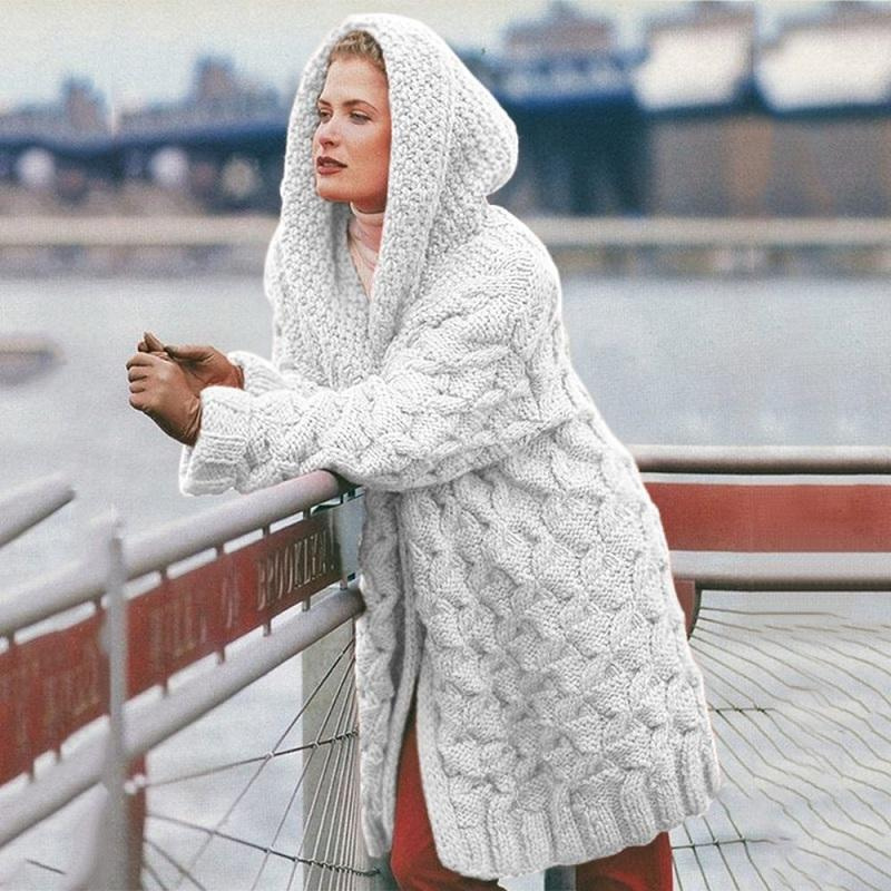 Femme en manteau à capuche en laine blanche, motif torsadé, appuyée sur une rambarde près d'un fleuve urbain. Mode hiver, style élégant.