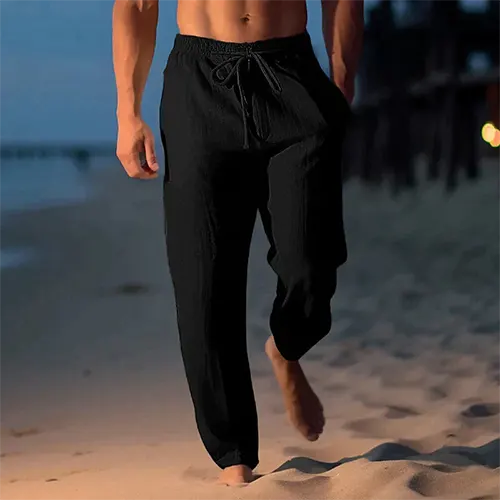 Homme marchant sur la plage au crépuscule, portant un pantalon noir décontracté. Mode masculine, style plage, confort estival.