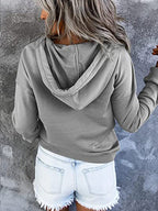 Femme en sweat à capuche gris, cheveux longs, shorts en jean déchiré, pose de dos contre un mur texturé. Mode décontractée, style urbain.