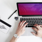 Ordinateur portable avec écran rose, mains tapant sur le clavier, clé USB insérée, carnet et stylo sur un bureau blanc.