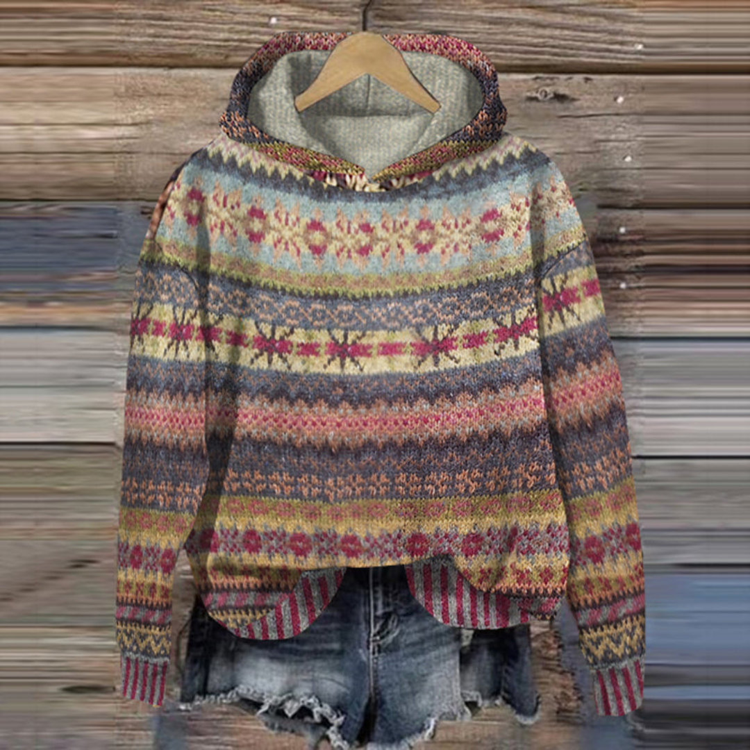 Pull-over à capuche en laine multicolore avec motifs nordiques suspendu sur un cintre en bois, devant un fond en bois rustique.