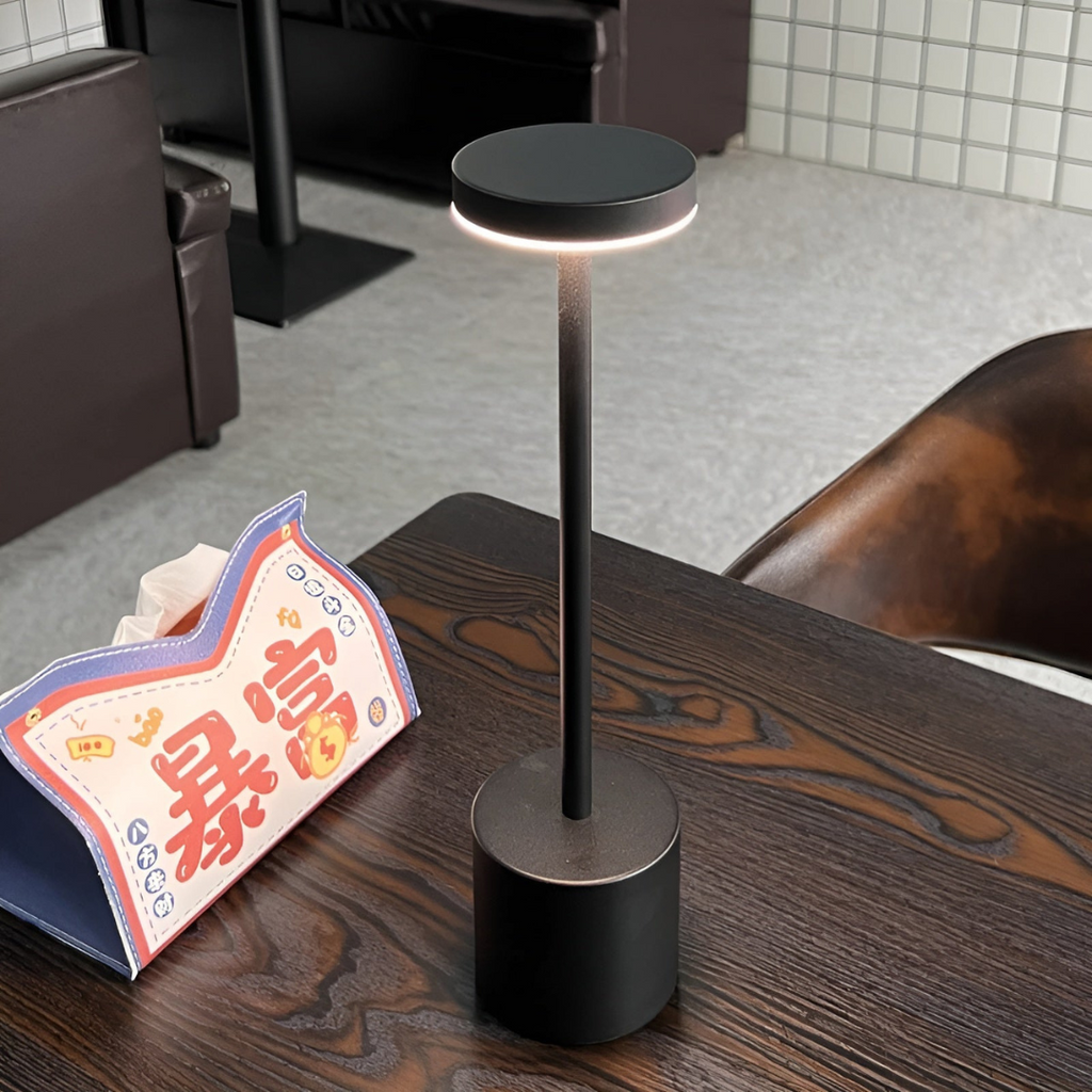 Lampe de table moderne noire sur une table en bois sombre, avec un menu en papier coloré à côté. Éclairage LED, design minimaliste, intérieur contemporain.