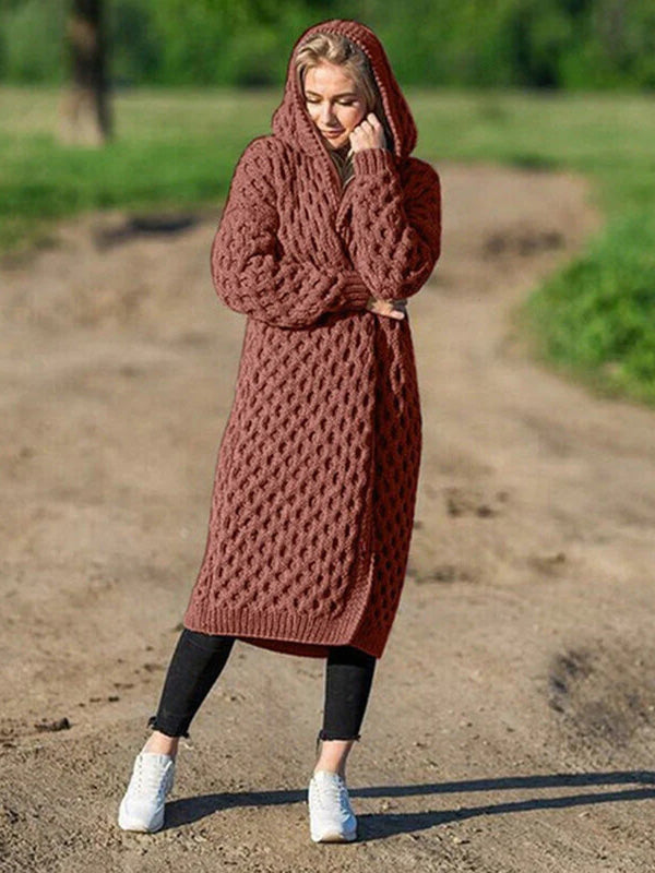 Femme portant un long cardigan en tricot marron avec capuche, debout sur un chemin de terre, style automne, mode décontractée, nature en arrière-plan.