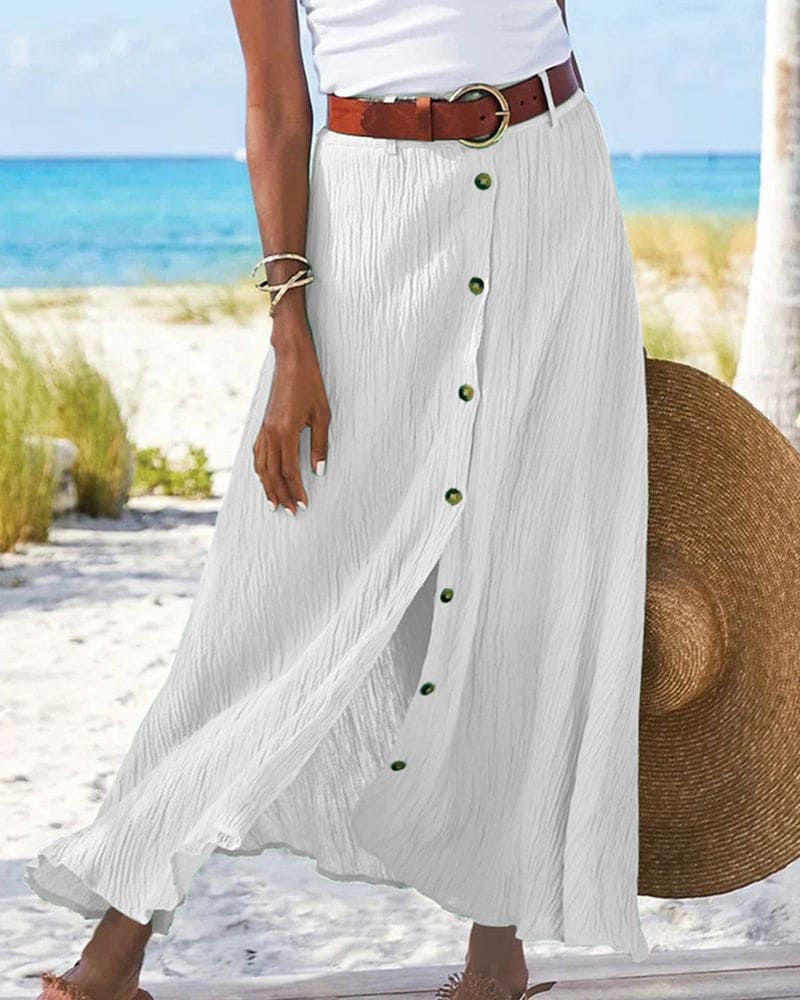 Femme en jupe longue blanche boutonnée, ceinture marron, tenant un chapeau de paille sur une plage ensoleillée avec mer bleue en arrière-plan.