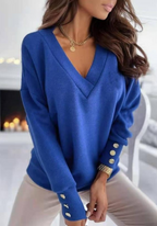 Femme portant un pull bleu à col en V, manches longues avec boutons dorés, posant dans un intérieur moderne. Mode féminine élégante et tendance.