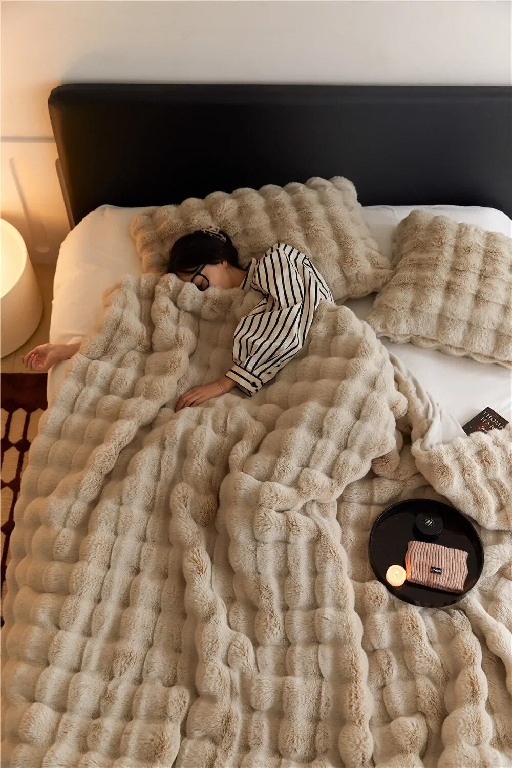 Femme dormant sur un lit avec couverture en fausse fourrure beige, coussins assortis, lampe de chevet allumée, ambiance cosy et relaxante.