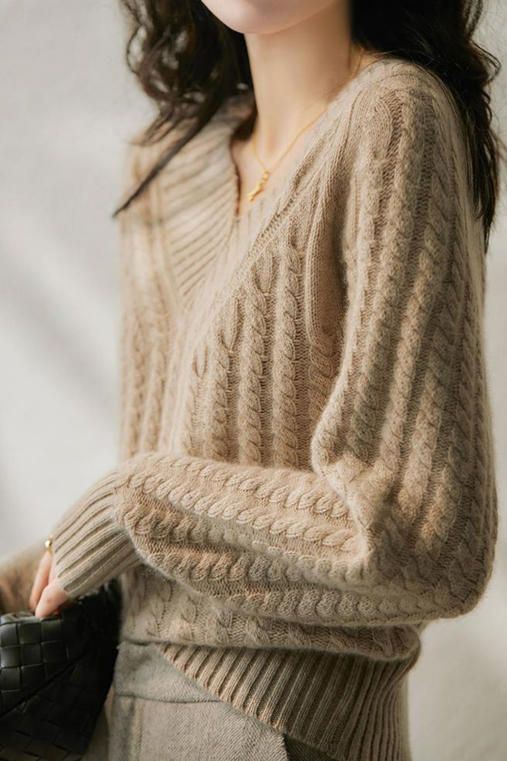 Femme portant un pull en laine torsadée beige, tenant un sac à main noir. Mode automne-hiver, style élégant et confortable.