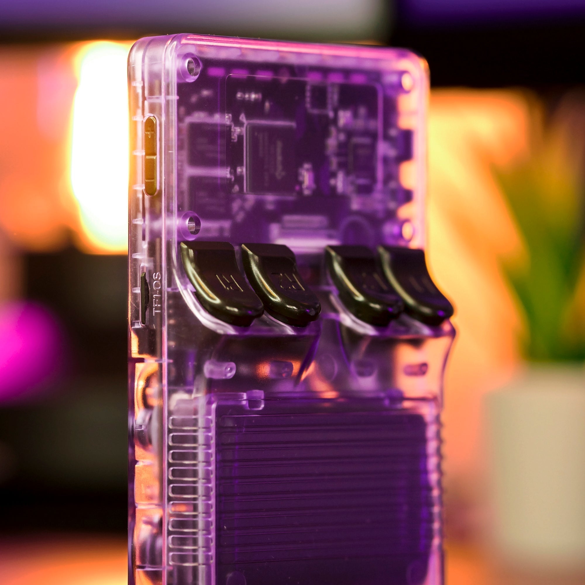 Console de jeu rétro transparente avec boutons noirs, circuits visibles, éclairage d'ambiance violet et orange, plante en arrière-plan.