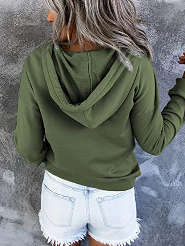 Femme de dos portant un sweat à capuche vert olive et un short en jean déchiré, posant devant un mur gris texturé. Mode décontractée tendance.