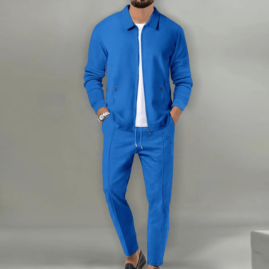 Homme portant un ensemble bleu élégant, veste et pantalon assortis, sur fond gris. Mode masculine, tenue décontractée chic, style moderne.