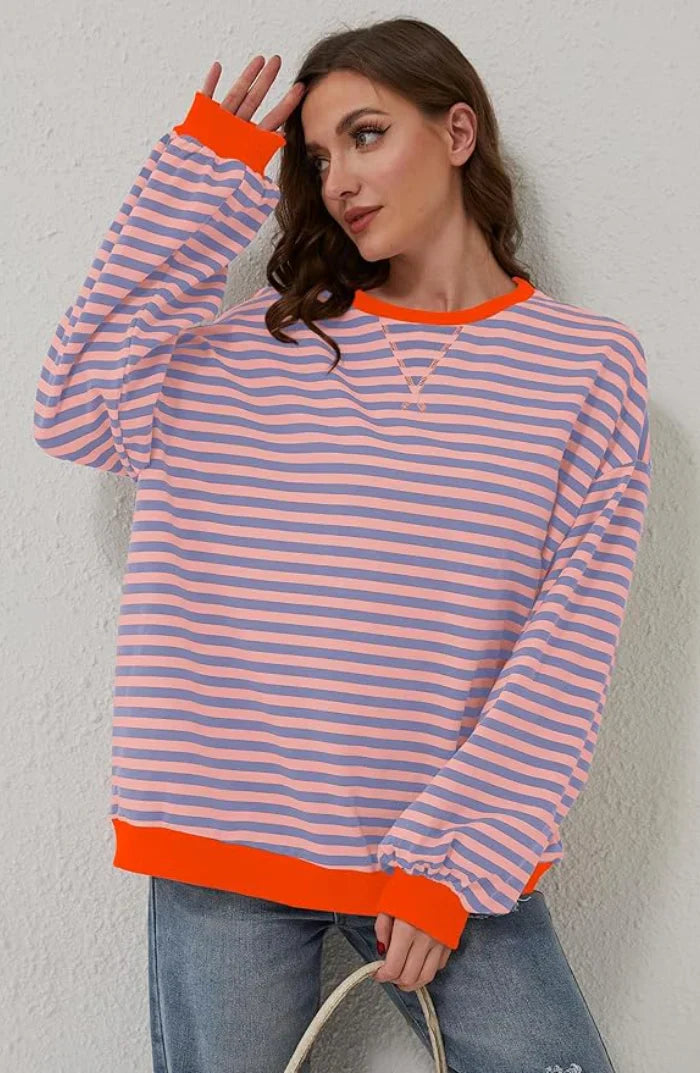Femme portant un pull rayé rose et violet avec des bordures orange, posant contre un mur blanc. Mode décontractée, tendance automne-hiver.