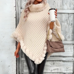 Femme portant un poncho en tricot crème avec bordure en fourrure, tenant un café à emporter et un sac à main beige, devant une porte en bois.