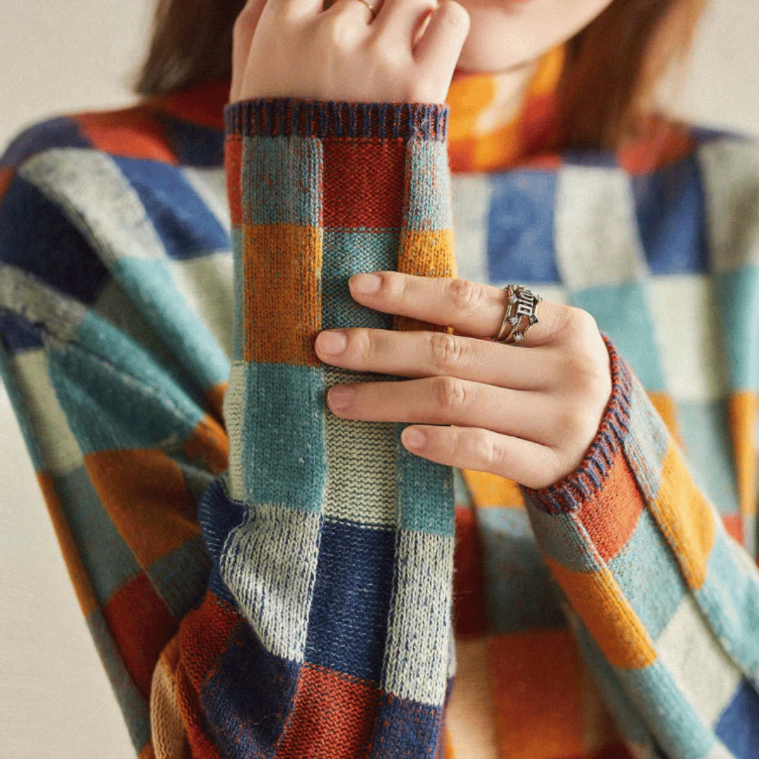 Pull à carreaux colorés en laine, manches longues, porté par une femme avec une bague argentée. Mode automne-hiver, style décontracté et tendance.