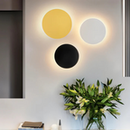Applique murale moderne avec trois cercles lumineux jaune, blanc et noir sur mur gris, accompagnée d'un vase de lys blancs. Décoration intérieure élégante.