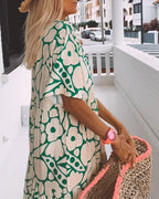 Femme en robe à motifs floraux verts, portant un chapeau de paille et un sac en osier, sur une terrasse blanche. Mode estivale, style bohème.