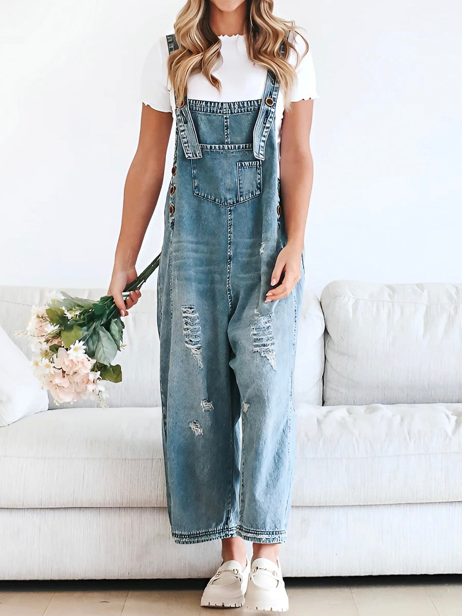 Femme en salopette en jean délavé, t-shirt blanc, tenant un bouquet de fleurs, posée devant un canapé blanc. Mode décontractée, tendance denim.