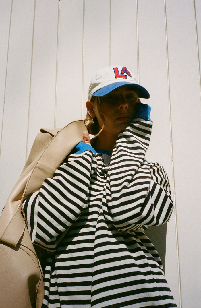 Jeune femme portant un pull rayé noir et blanc, casquette LA blanche et bleue, sac beige, posant devant un mur blanc ensoleillé.