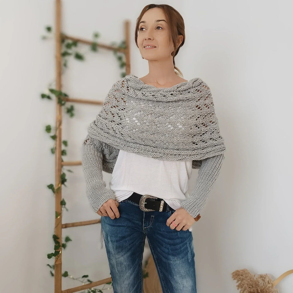 Femme portant un châle en tricot gris sur un pull blanc, posant devant une échelle en bois décorée de plantes vertes. Mode automne-hiver décontractée.