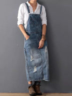 Femme portant une robe salopette en jean déchiré, chemise blanche, bottines marron, posant contre un mur gris. Mode décontractée, tendance denim.