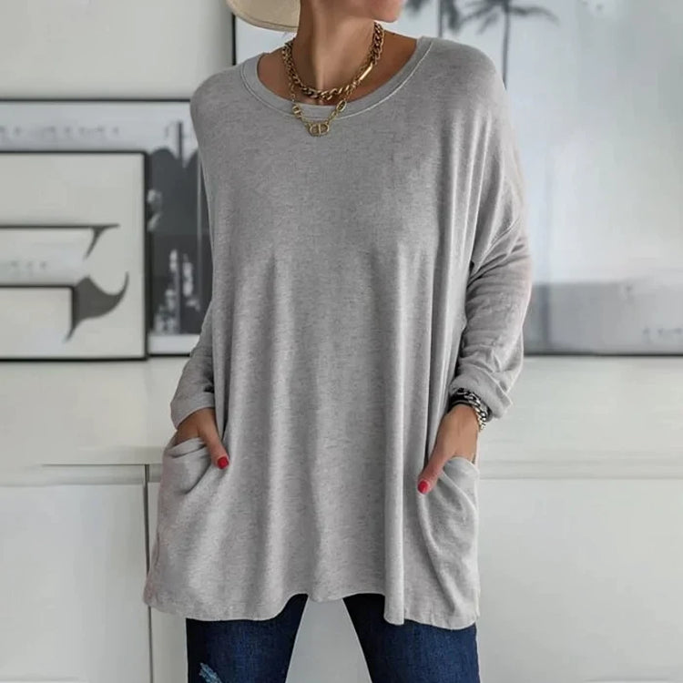 Femme portant un pull gris ample avec poches, collier doré, et jeans bleus, posant devant un mur décoré. Mode décontractée élégante.