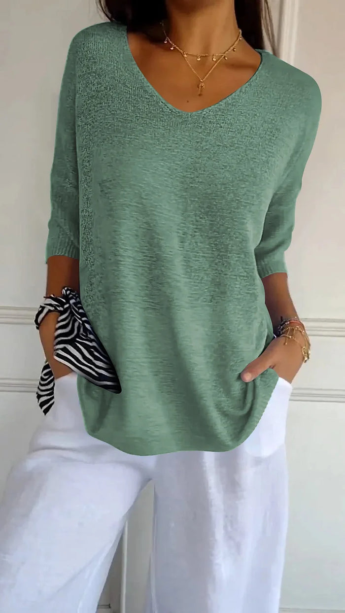 Femme portant un pull vert à col en V, manches trois-quarts, avec un pantalon blanc. Accessoires: collier doré, bracelet, foulard rayé noir et blanc.