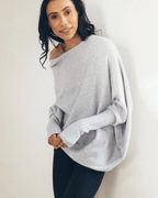 Femme portant un pull gris ample, manches longues, col bateau, posant contre un mur blanc. Mode décontractée, tendance automne-hiver.