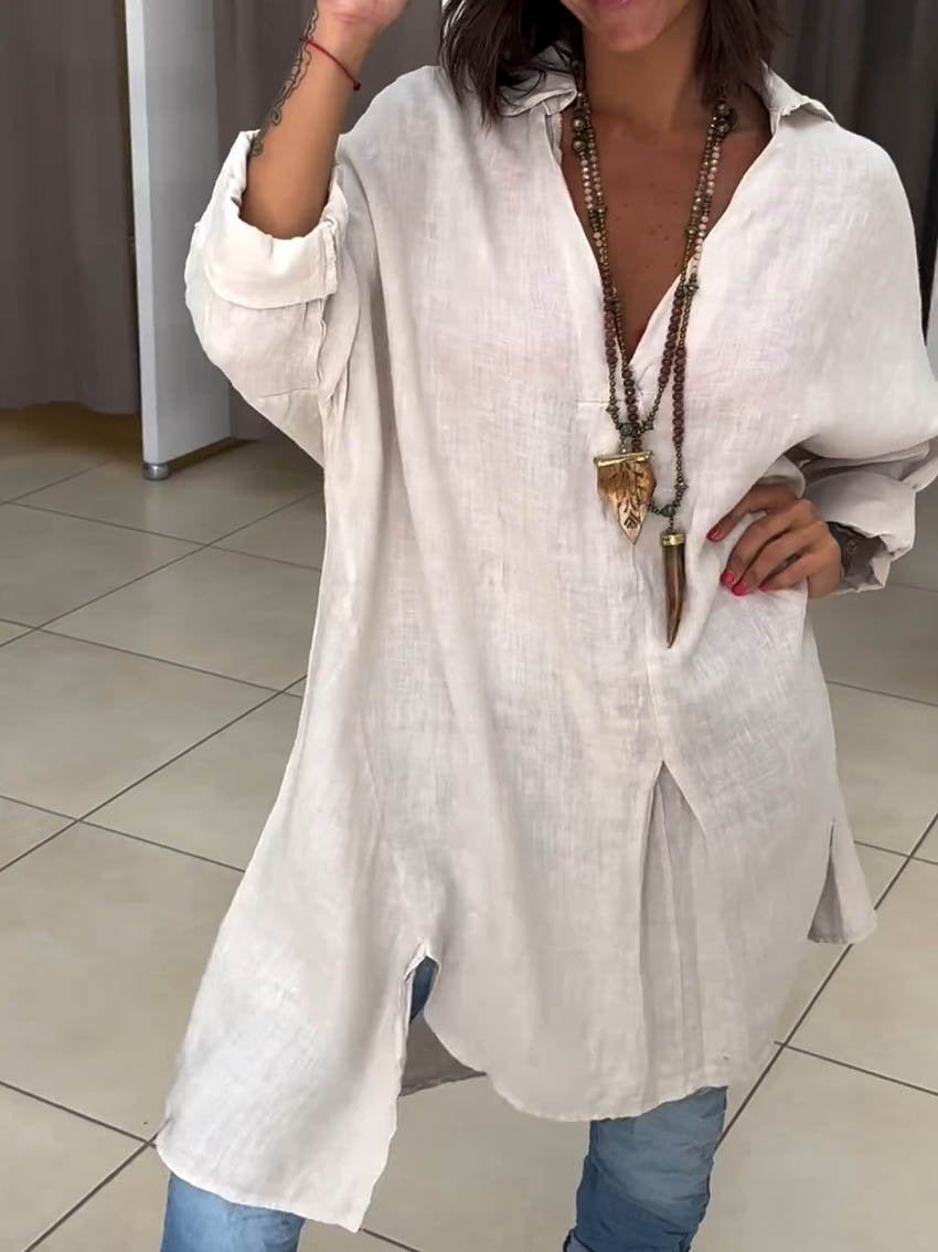 Femme portant une tunique blanche en lin, col ouvert, manches longues, avec collier ethnique, posant sur un sol carrelé. Mode décontractée chic.