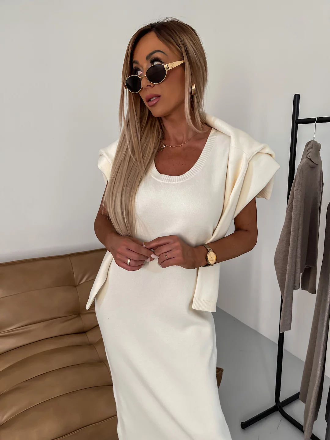 Femme élégante en robe blanche, lunettes de soleil, posant près d'un canapé beige et portant un pull sur les épaules. Mode chic et moderne.