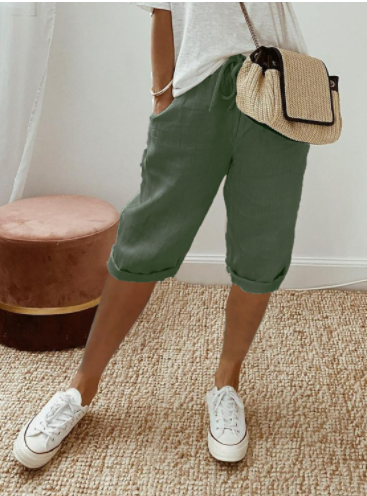 Femme portant un short vert en lin, t-shirt blanc, baskets blanches et sac en osier, posant dans une pièce moderne avec pouf rose.