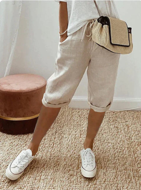 Femme portant un short beige en lin, t-shirt blanc, baskets blanches et sac à bandoulière en paille, debout sur un tapis en jute.